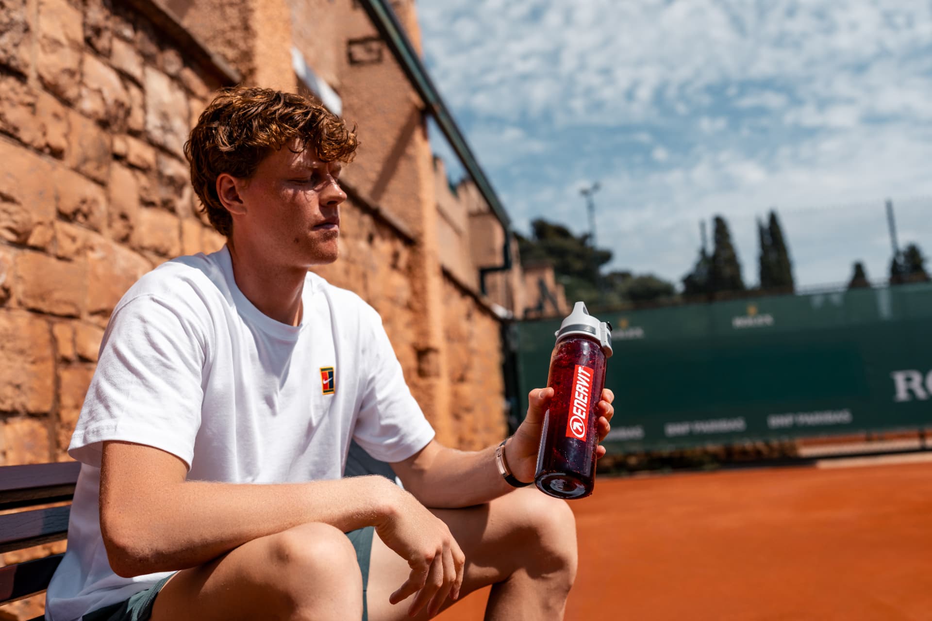Tennis player resting on a clay court bench holding an Enervit bottle